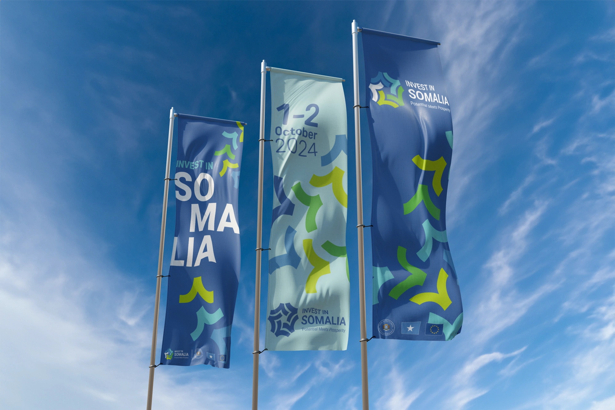 Three vertical corporate banners waving against a blue sky with brand patterns and the event dates.